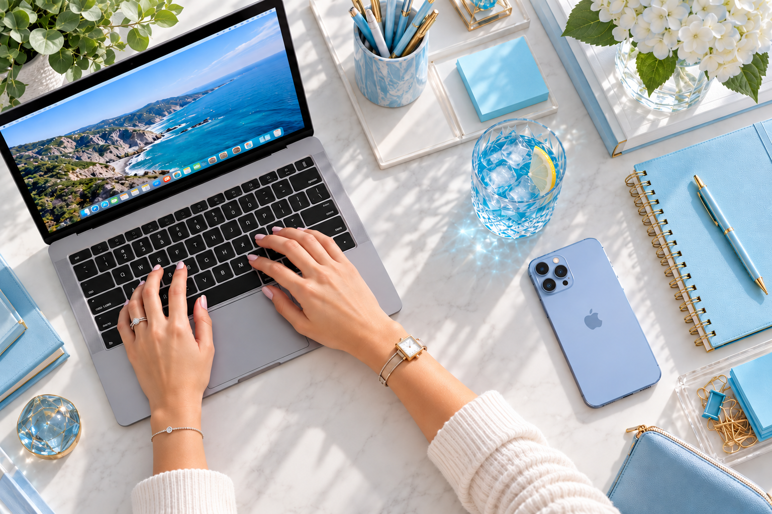Overhead view of hands typing on a laptop surrounded by blue office supplies and a crystal glass of iced blue drink